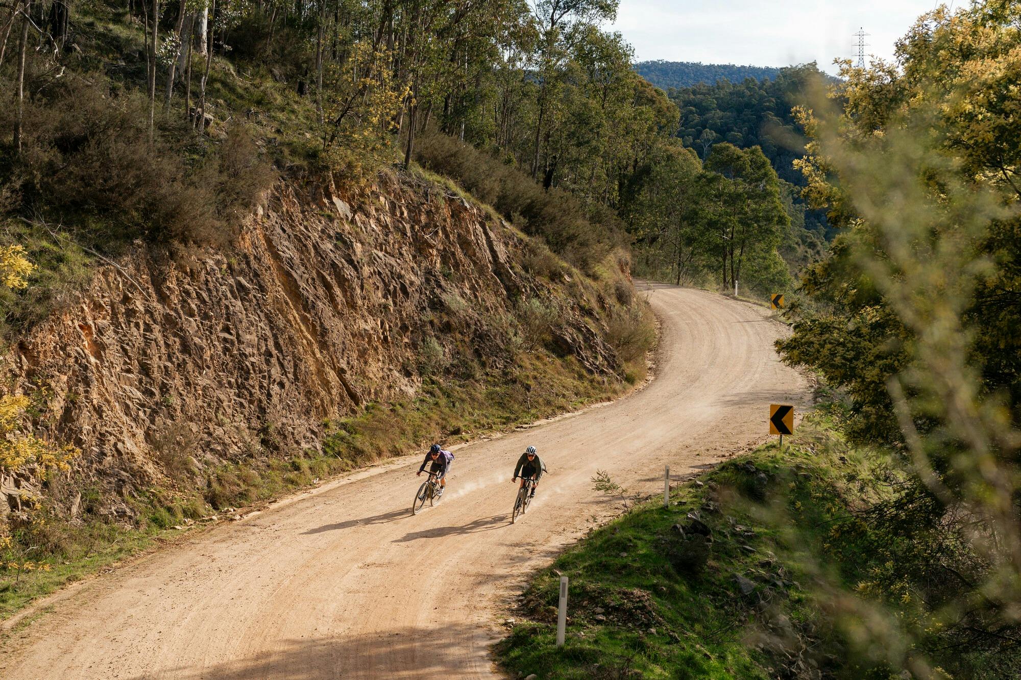 Two cyclists riding on a gravel road cut into the side of a hill, lush green surrounds and sunny day