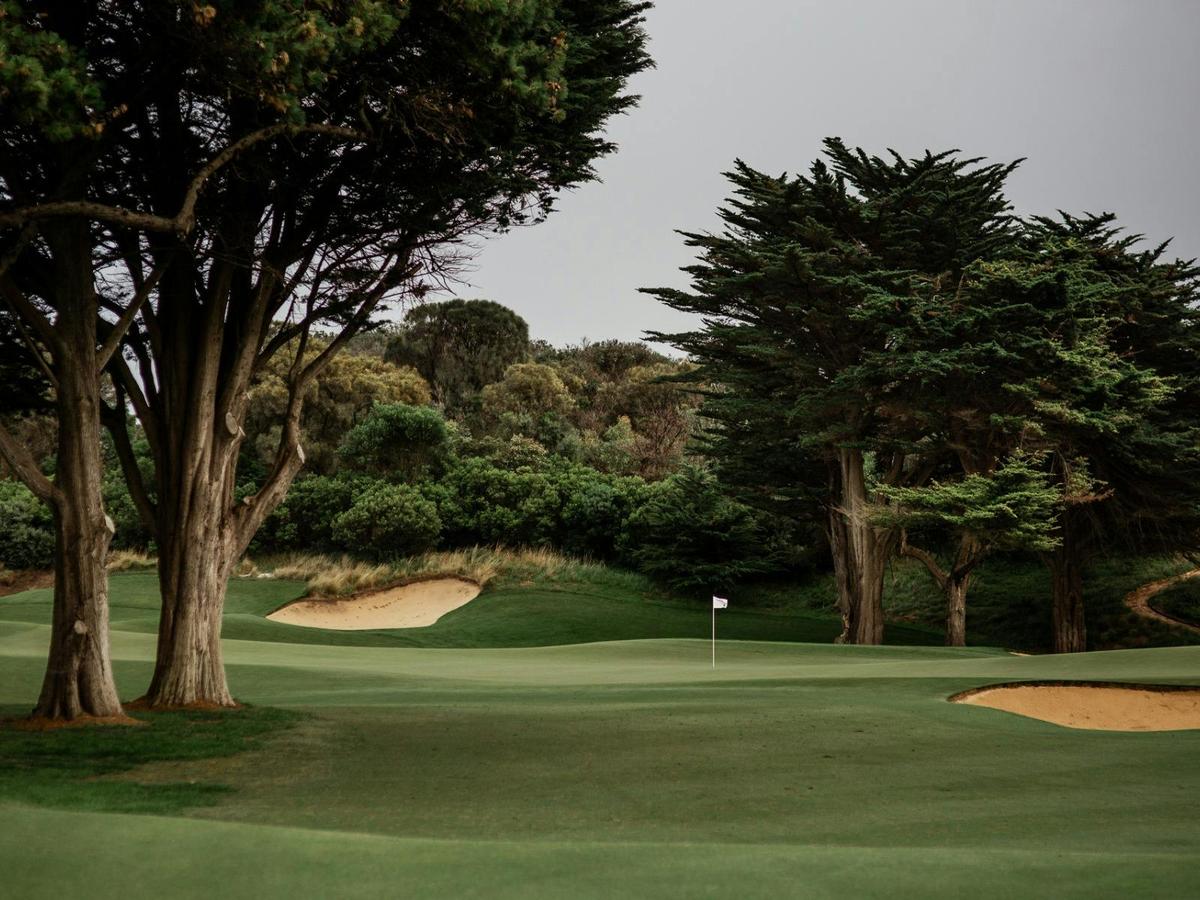 Golf hole framed by tall trees, pristine bunkers, and green under a soft, cloudy sky.