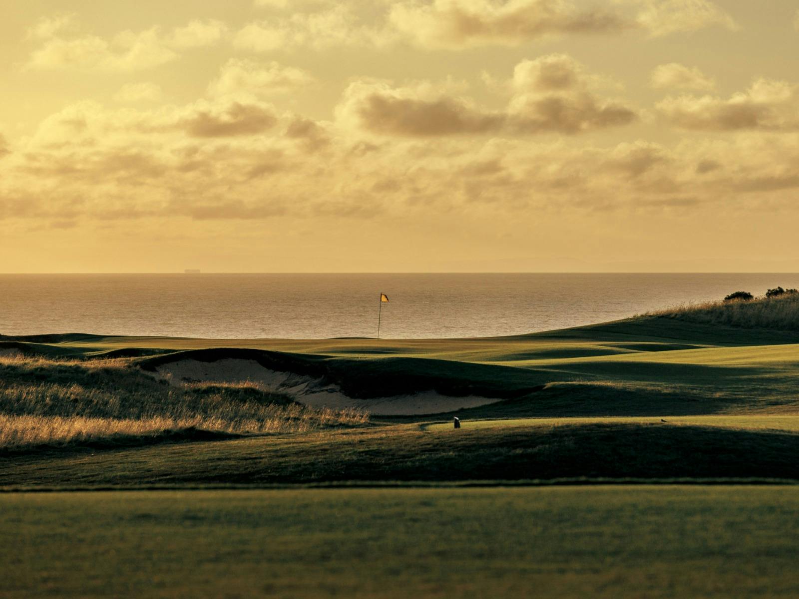 Golf hole with flagstick, green fairways, and ocean horizon under a warm golden sunset sky