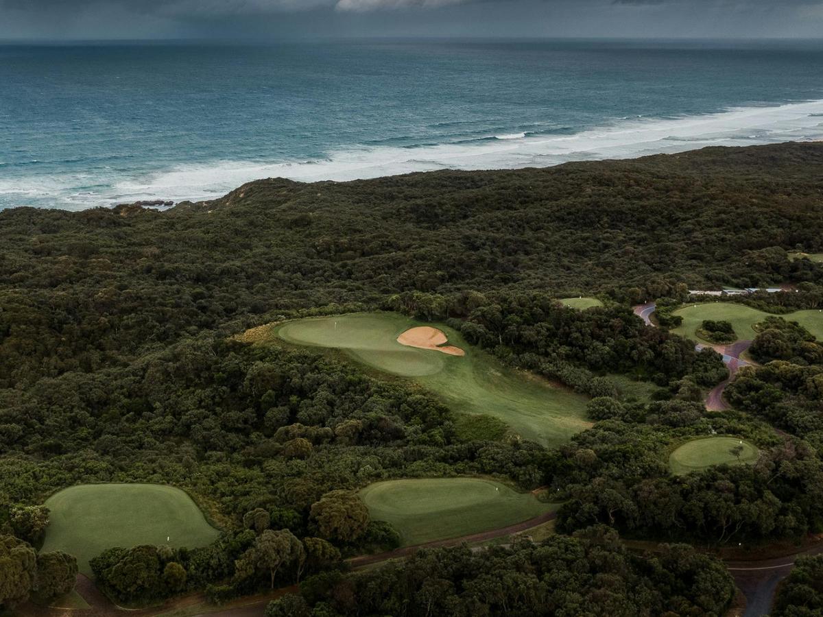 Aerial view of a golf course with fairways, bunkers, greenery, and ocean waves in the background