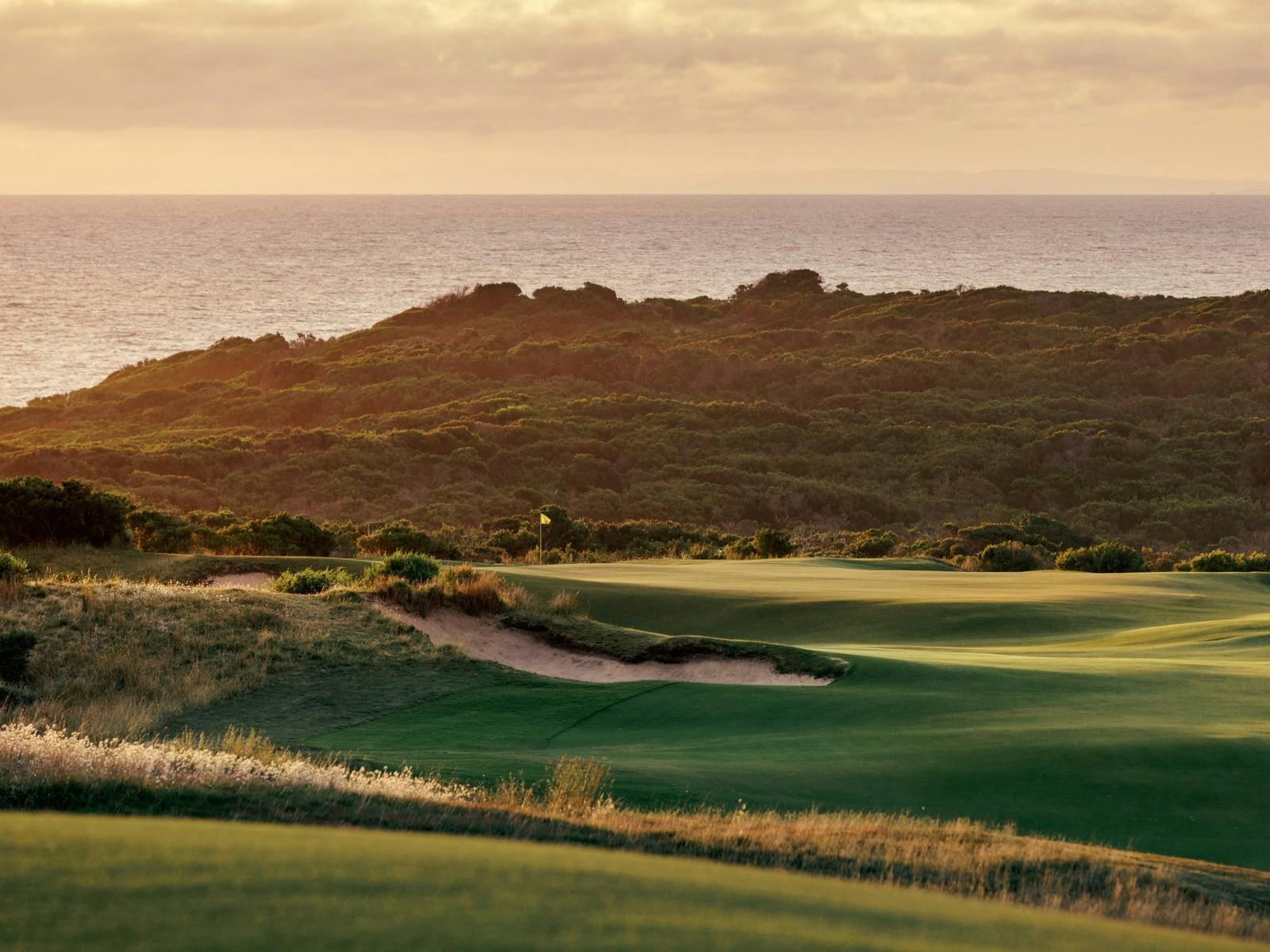 Sunset view of a golf course with fairways, bunker, coastal greenery, and ocean in the background.