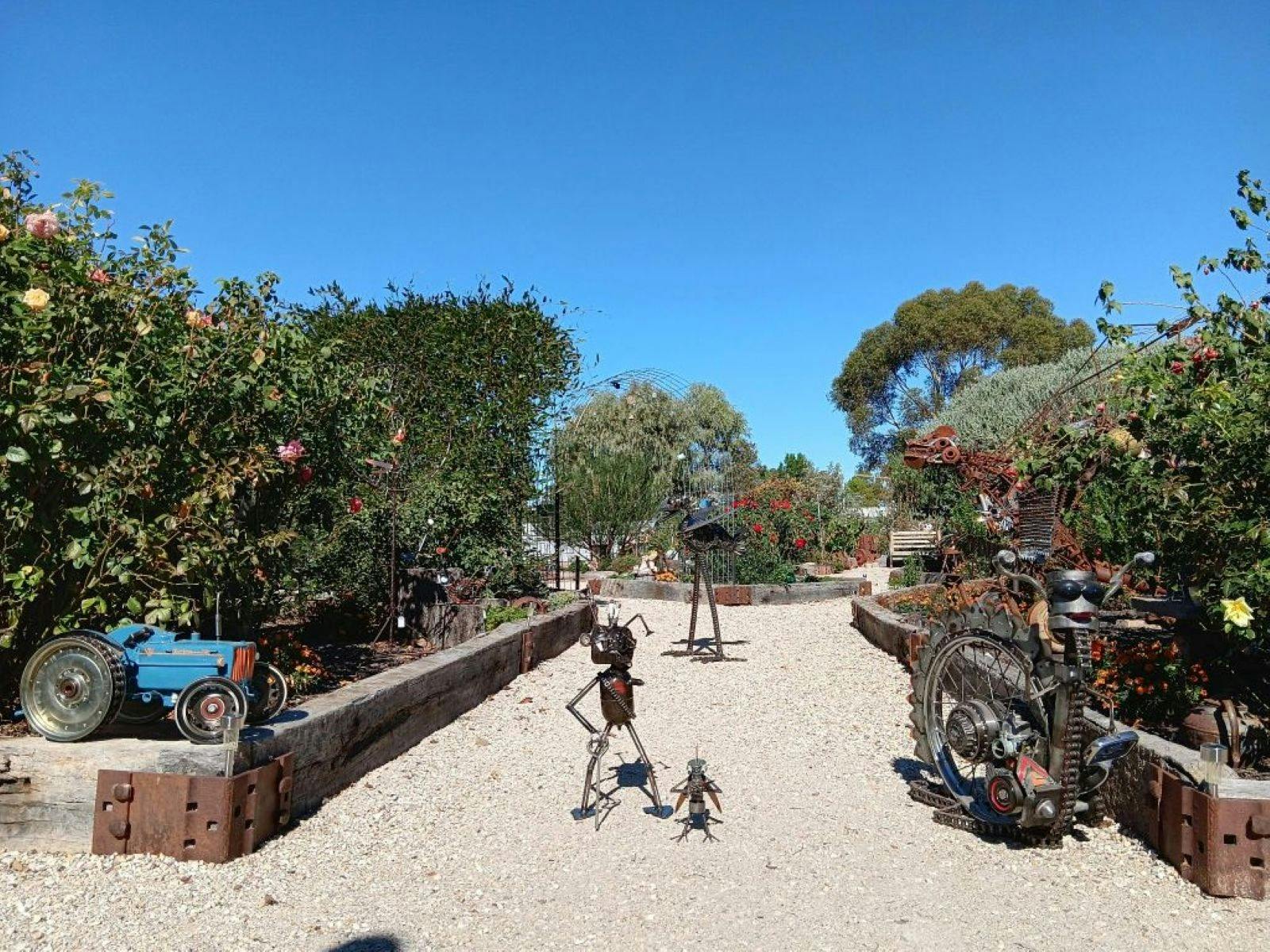 Wide gravel pathway through raised garden beds and a metal sculpture in the centre of the path