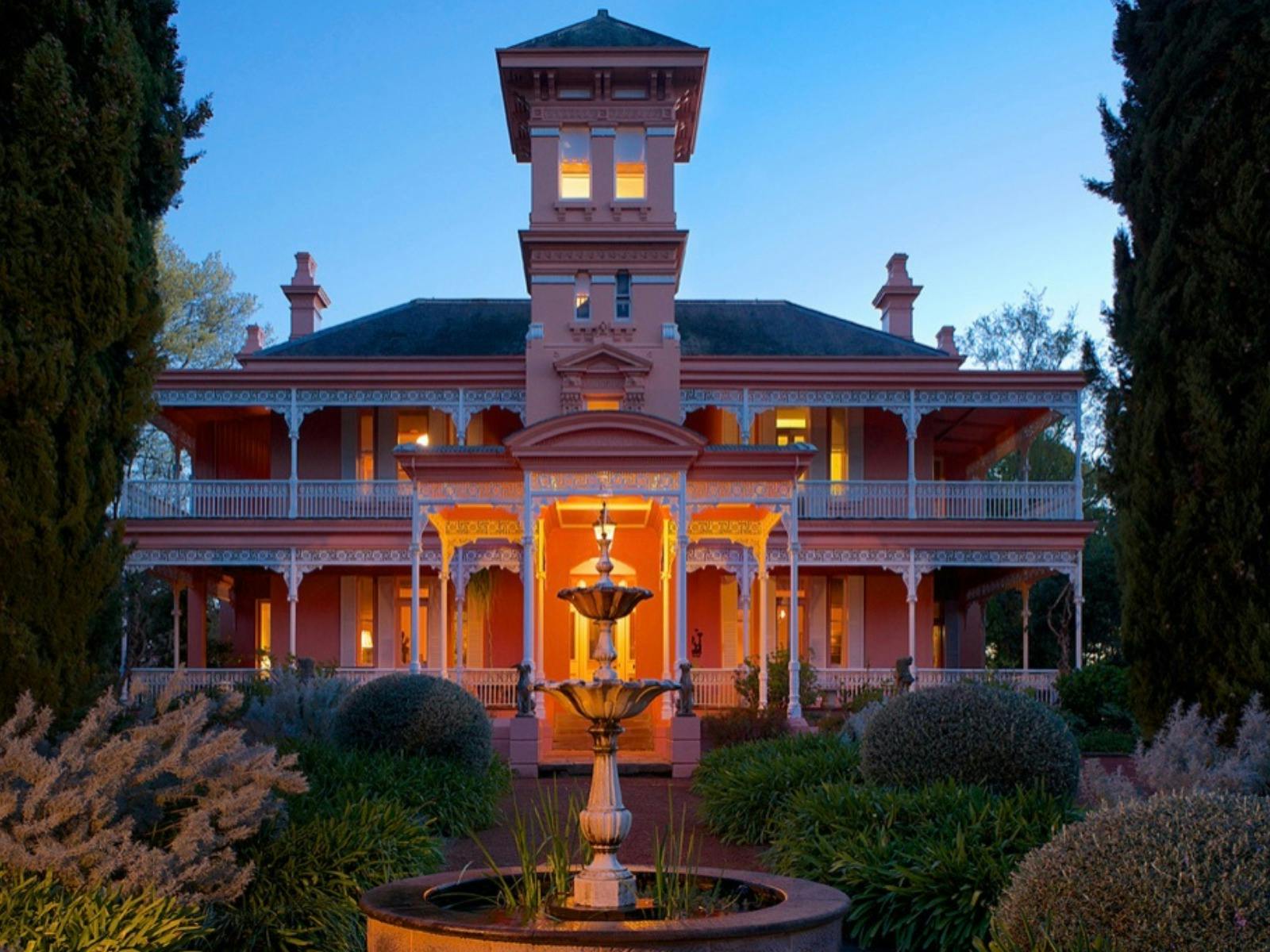Lights emit from a pink mansion surrounded by lush gardens and a fountain out front