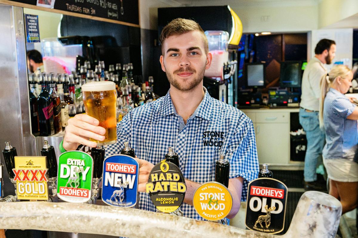 Bartender in checkered shirt holds a fresh pint behind beer taps.