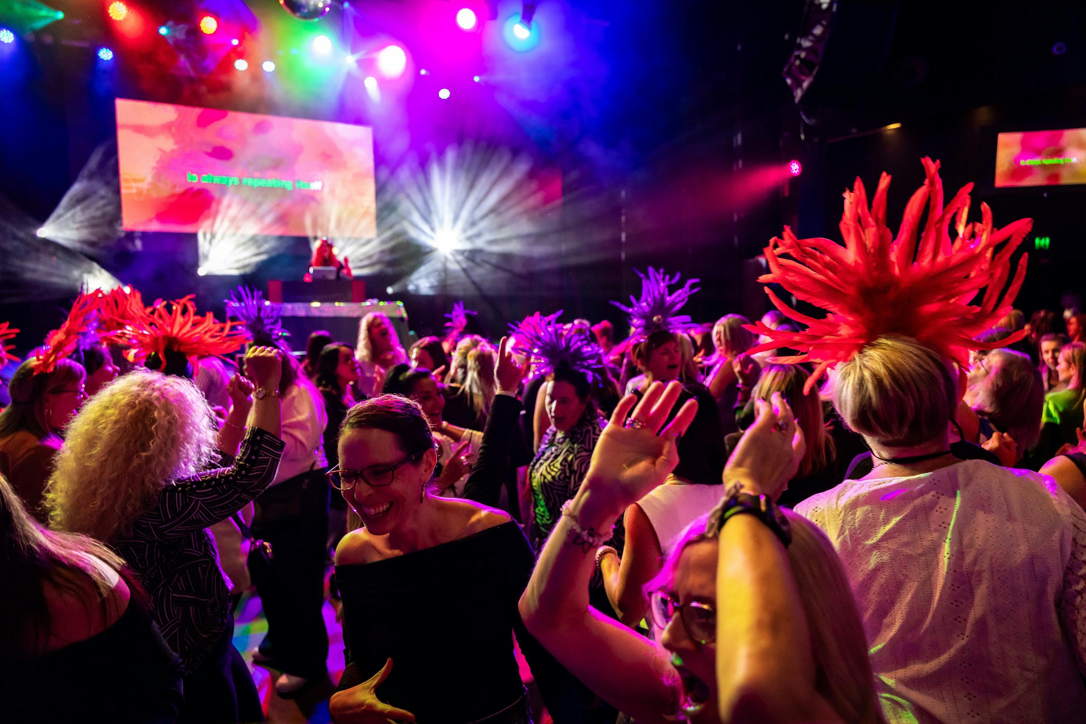 A crowd of ladies dance in front of the stage and bright stage lights, some wear feathered headbands