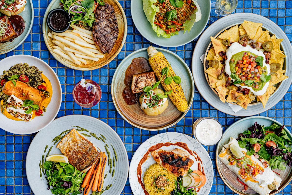 Flat lay of colorful dishes including steak, nachos, fish, and cocktails on a blue tile table.