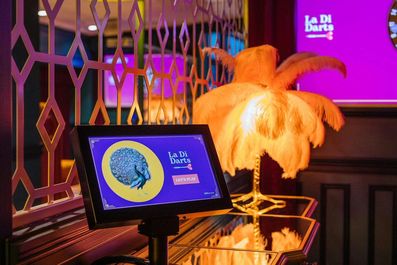Screen displays La Di Darts logo. Decorative lamp on table.