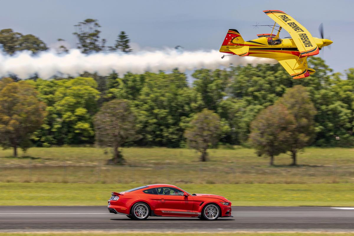 The Wolf Pitts Pro biplane makes a low-level pass above a red Shelby Mustang on the runway