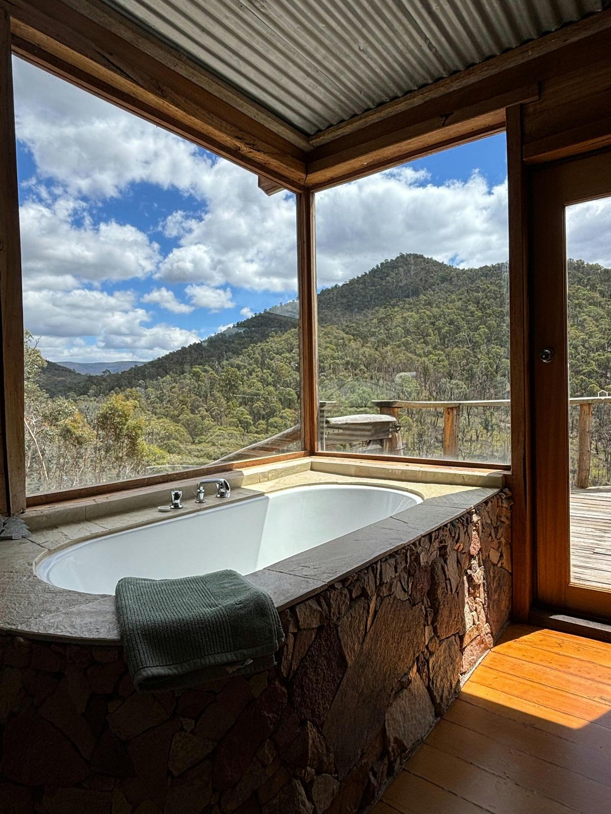 A stone tiled bathtub with floor to ceiling window that frame a mountainous bush landscape.