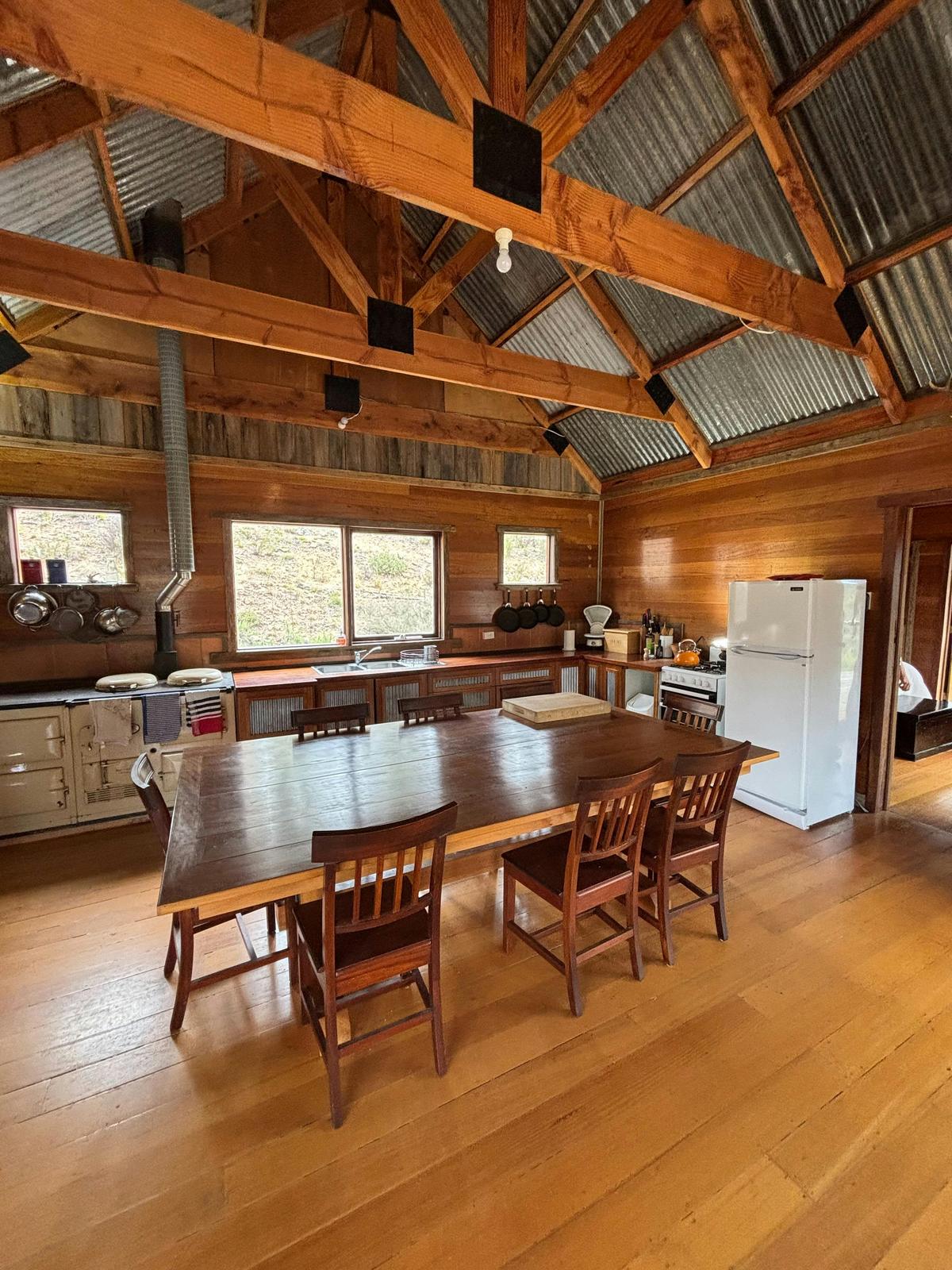 A large bespoke wooden dining table framed by well stocked, rustic open plan kitchen.