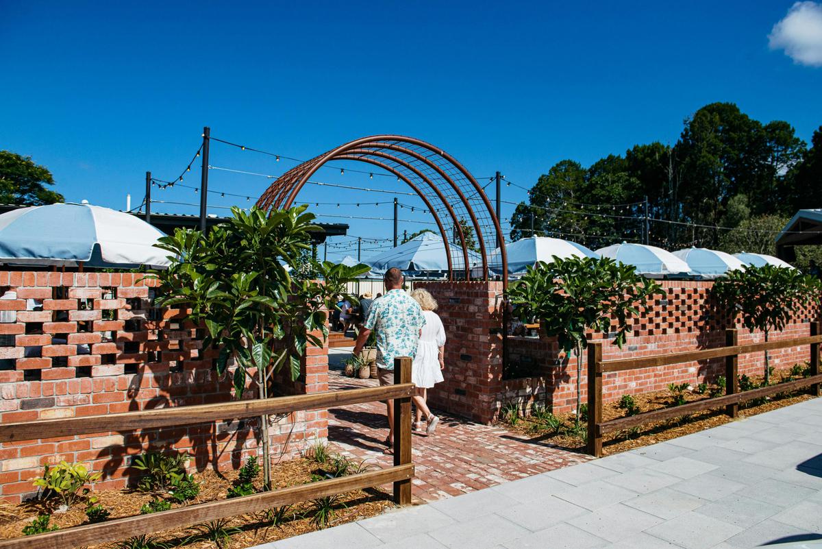 A couple walks through a brick archway entrance into a sunlit outdoor patio area with umbrellas