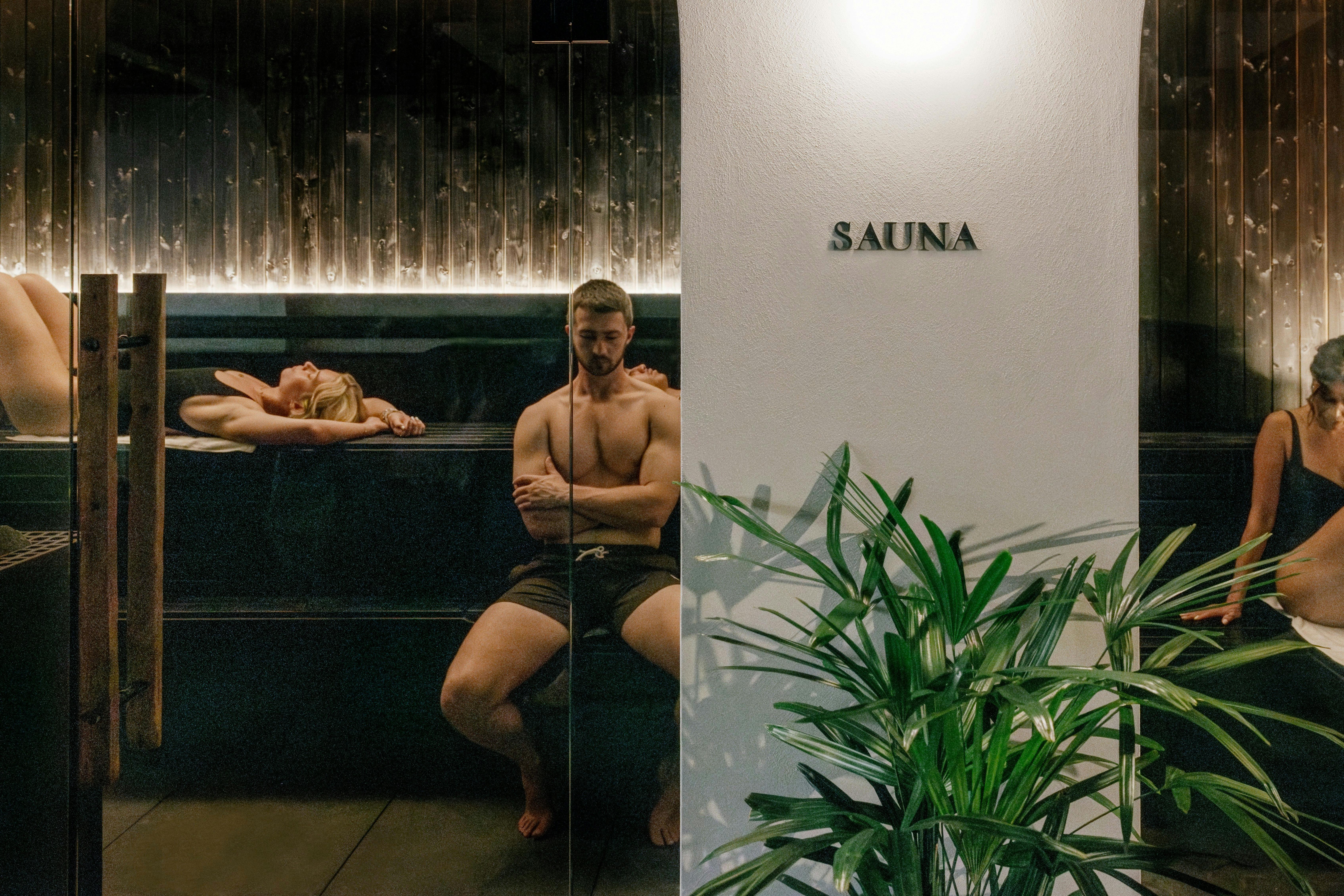 A group of people relaxing in a black timber sauna, with arches, plants and a sauna sign in front