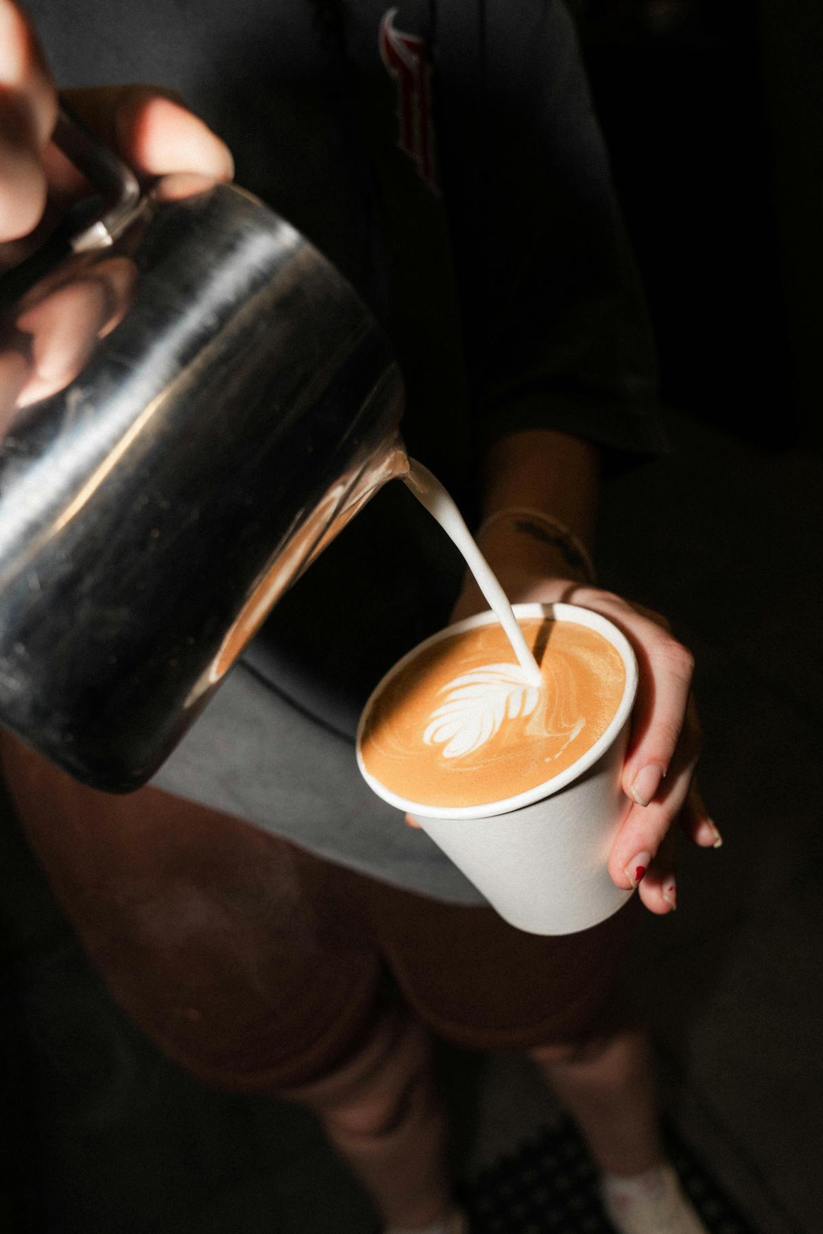 A white take away cup with milk being poured into it almost full with latte art