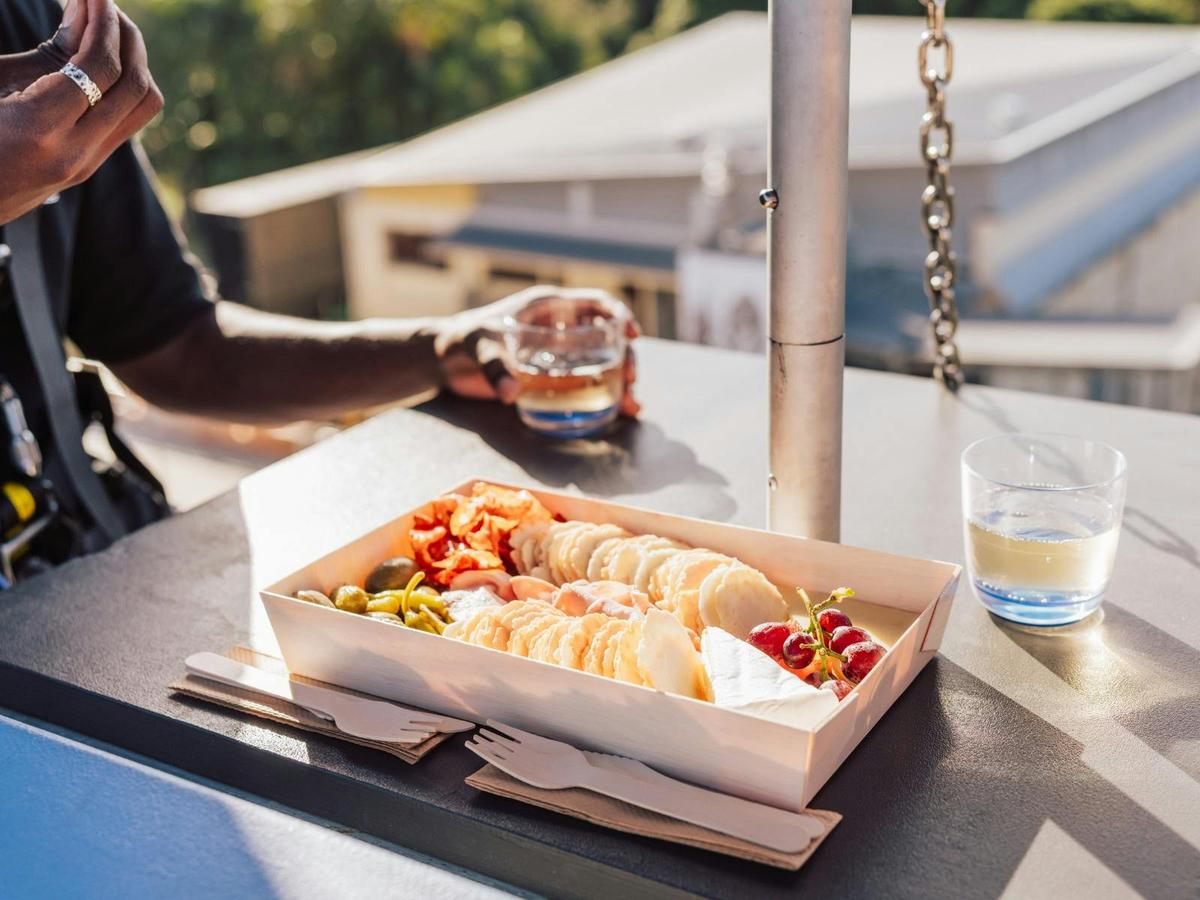 A box with assorted crackers, cheese, grapes, and olives on a table under sunlight.