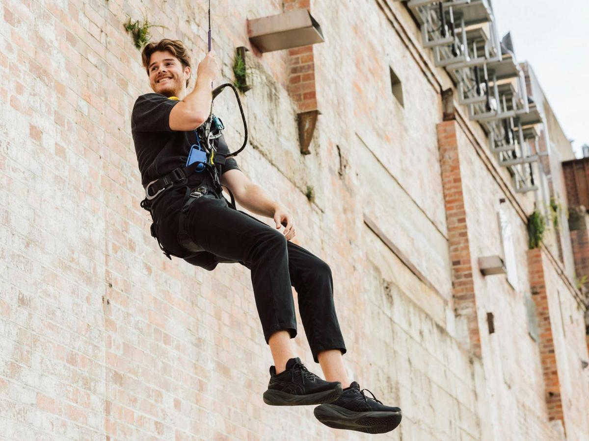 A person in a harness and black clothing smiles while descending a brick wall on a dropline.