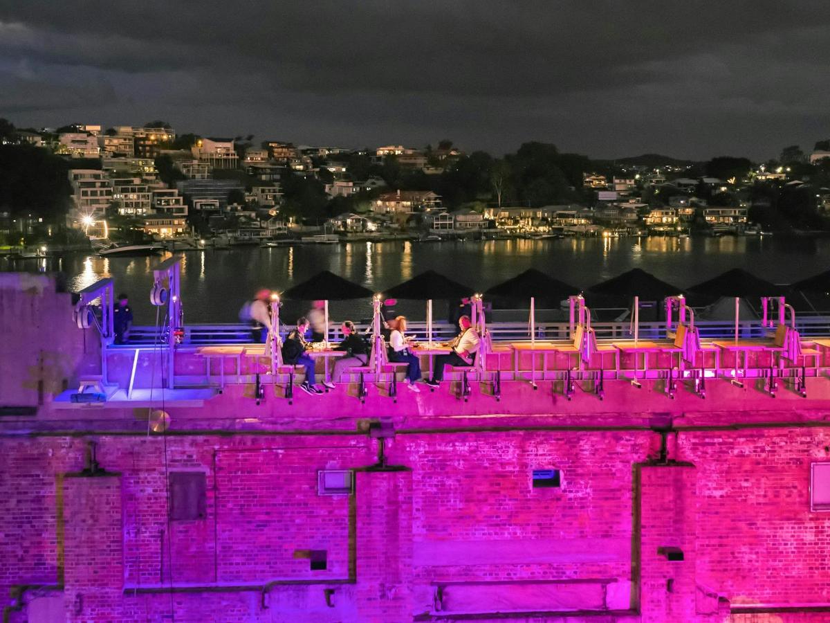 People dining on a rooftop above a lit pink brick building at night, overlooking a river.
