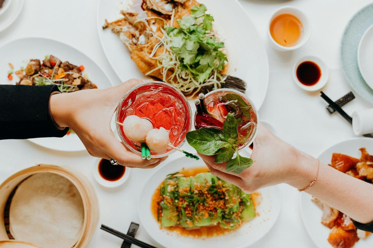 Two people hold two drinks above a spread of food.