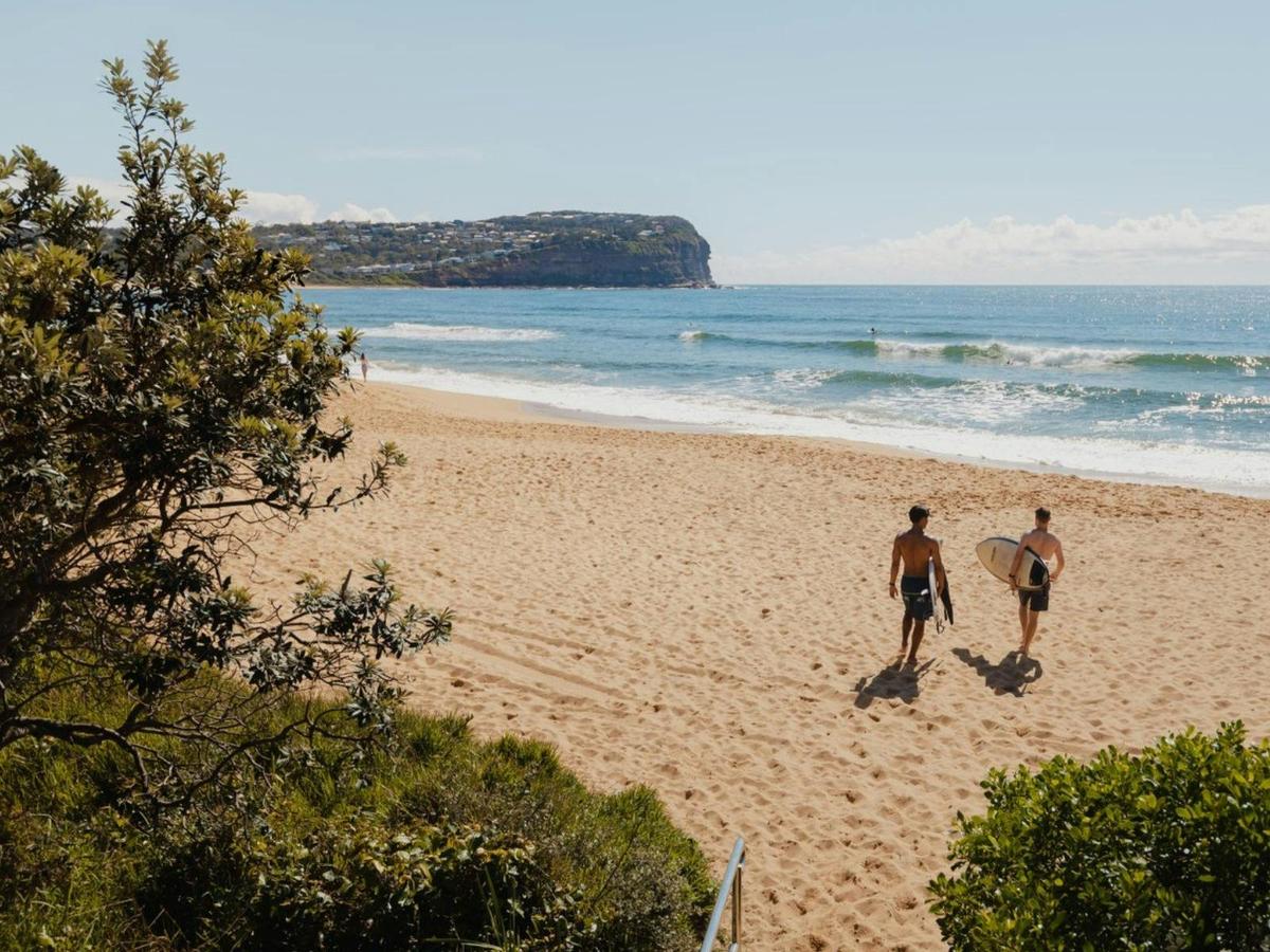 Two surfers walking across a sandy beach towards the ocean, with a coastal headland in the backgroun