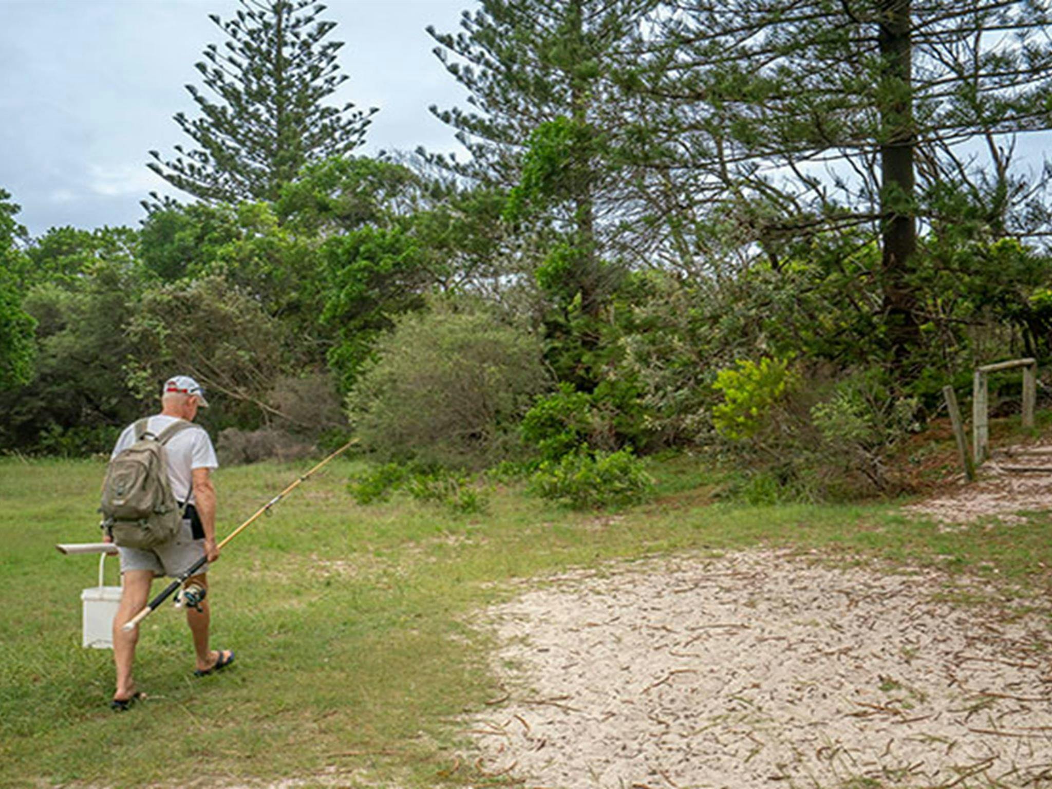 A man with a fishing rod walks across the grass toward a sandy trail and small set of steps. Photo