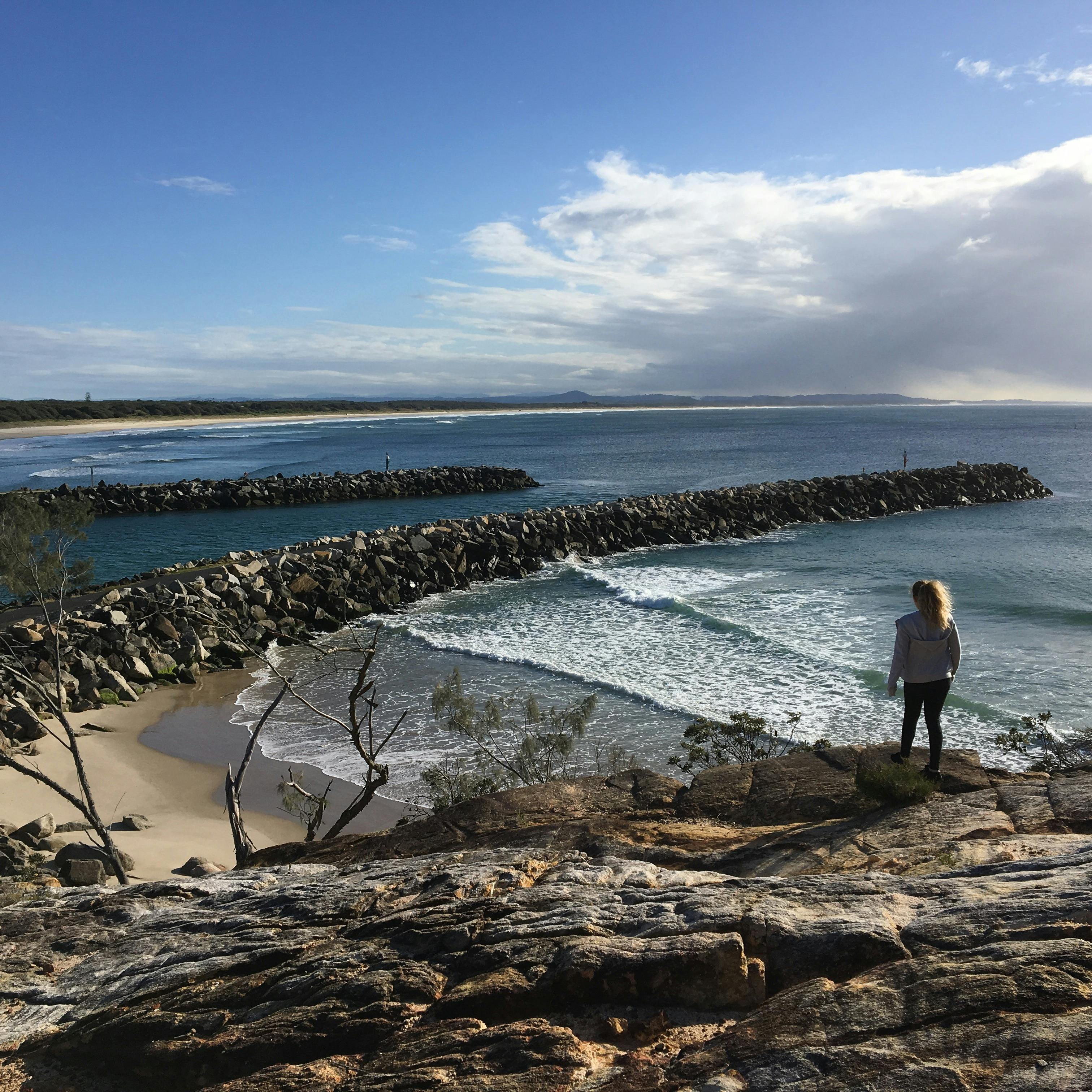 Girl standing on a rock looking down at Shark Bay. Evans Head River and wall in the background.