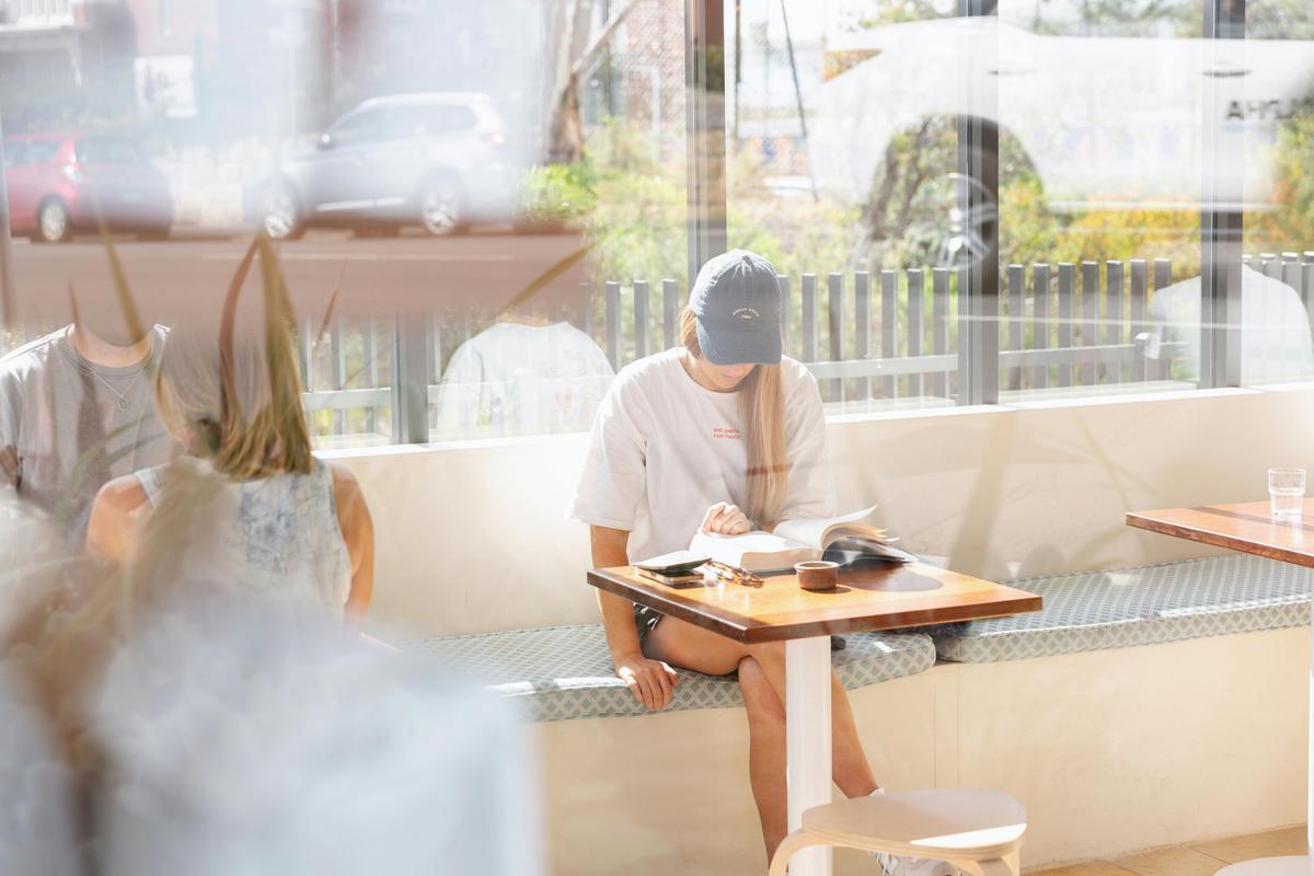 A woman reads a book in the sunshine at a table inside Abercrombie Coffee.
