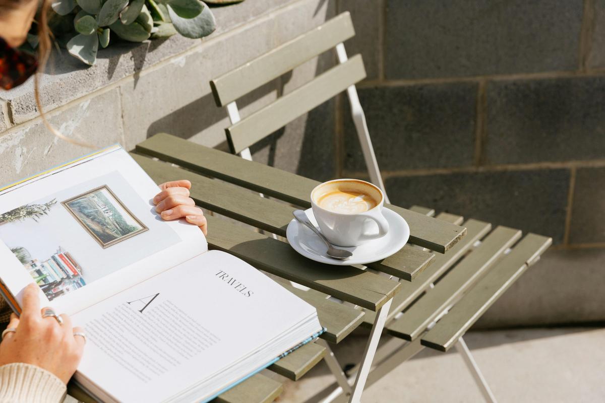 An open book and a partially drunk coffee are drenched in sunshine at an outside table.