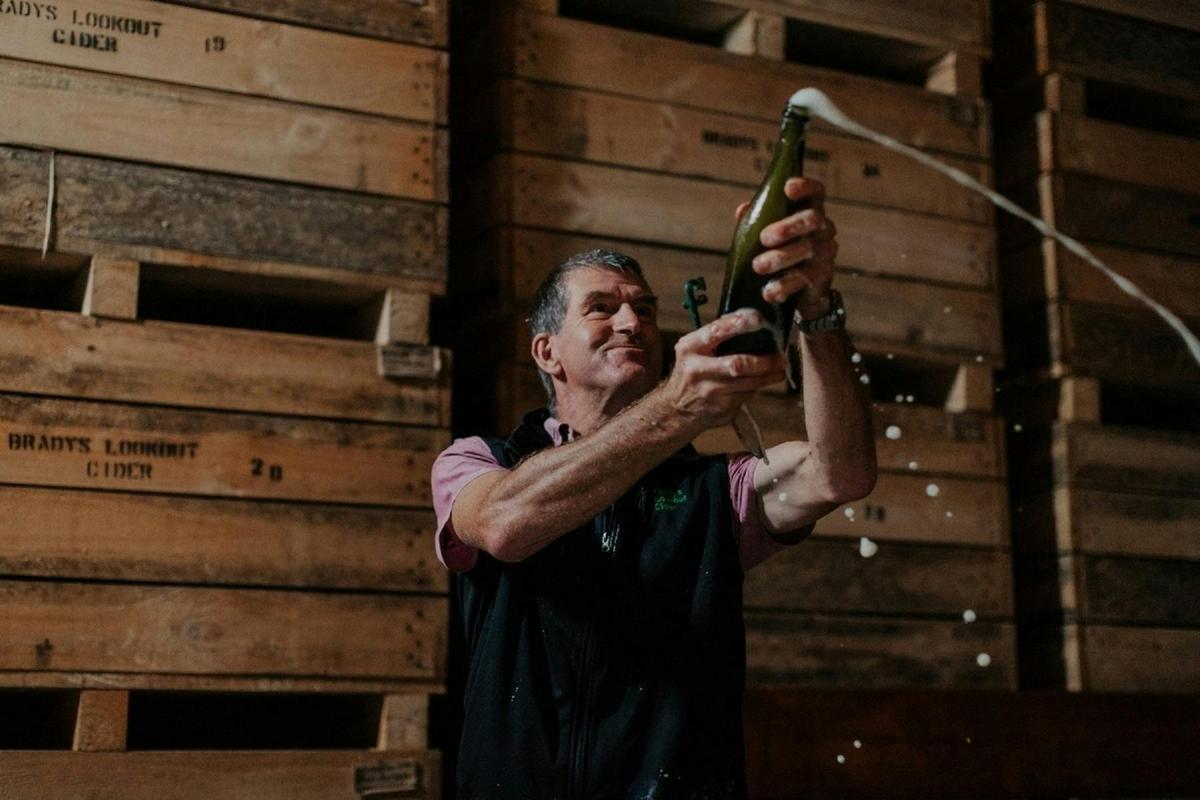 Bradys Lookout Cider. A man holding a bottle of wine up high as it spurts out onto the ground.