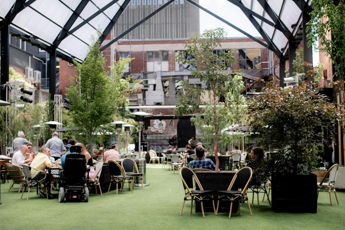Tables sit on green lawn, surrounded by chairs, potted trees and people under the Cathedral roof