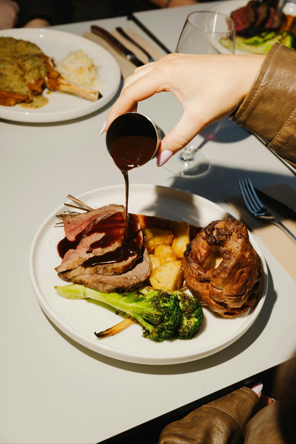 Gravy being poured over a Sunday roast with sliced lamb, roast potatoes, broccoli, and a Yorkshire