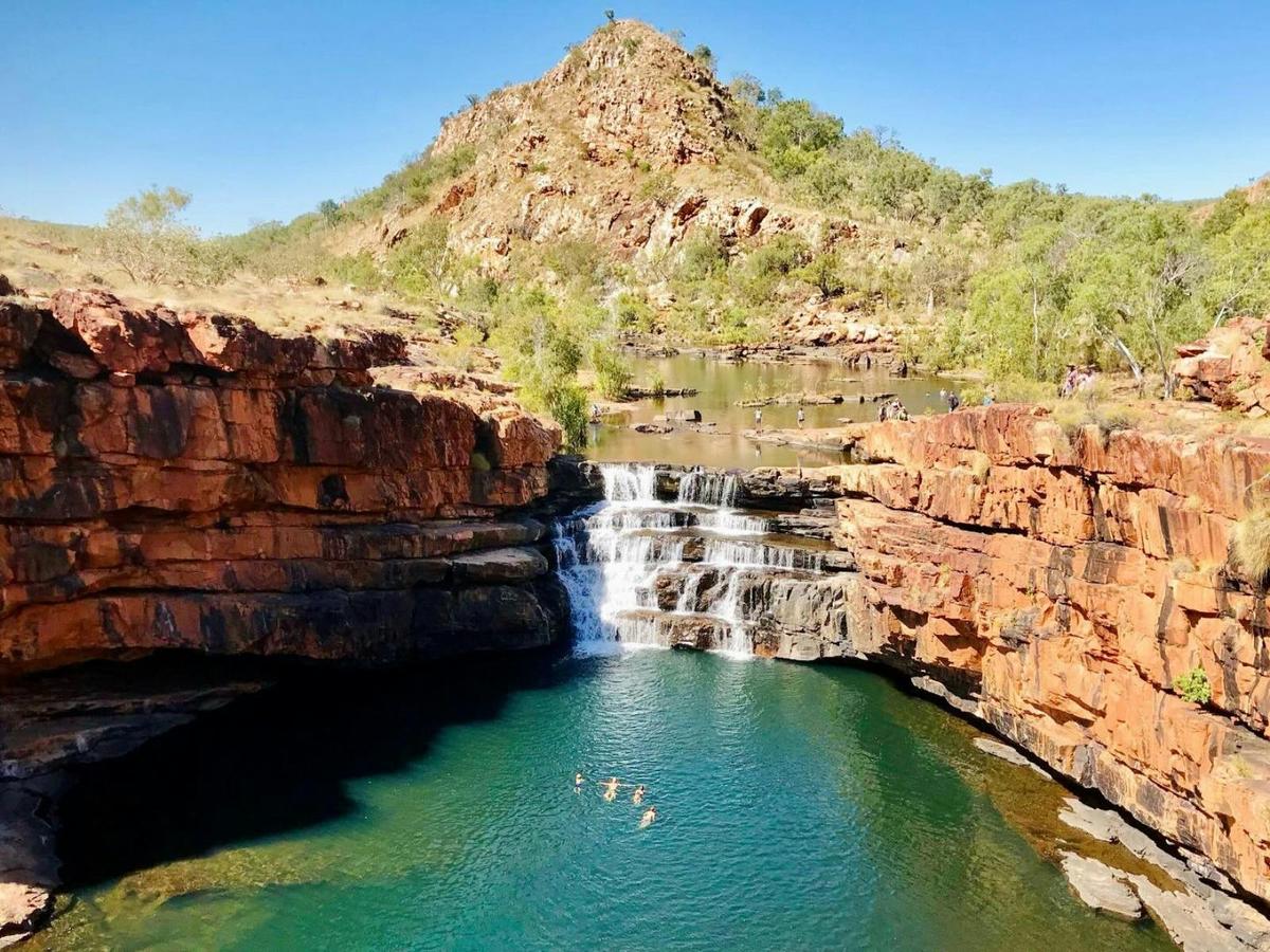 Turquoise pool beneath a small waterfall, framed by red rock cliffs and bushland.
