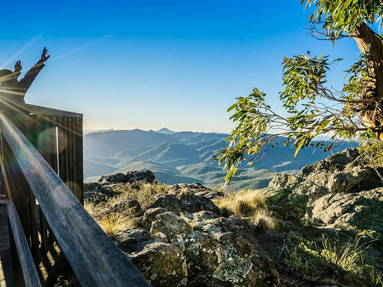 A visitor throws their hands in the air while standing on Governor lookout, with a vista of