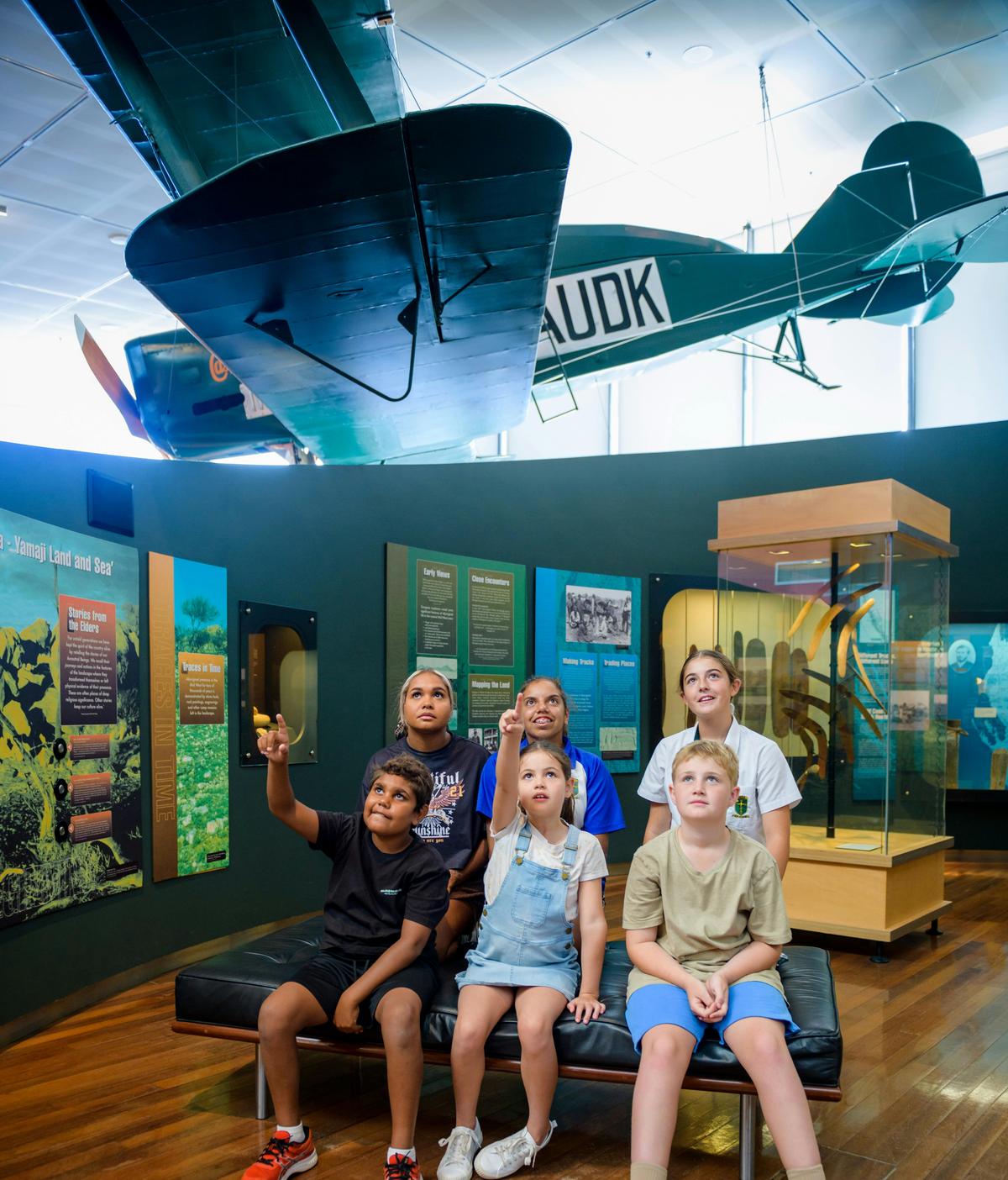 a group of children look up to an aviation display with panels of information in the background