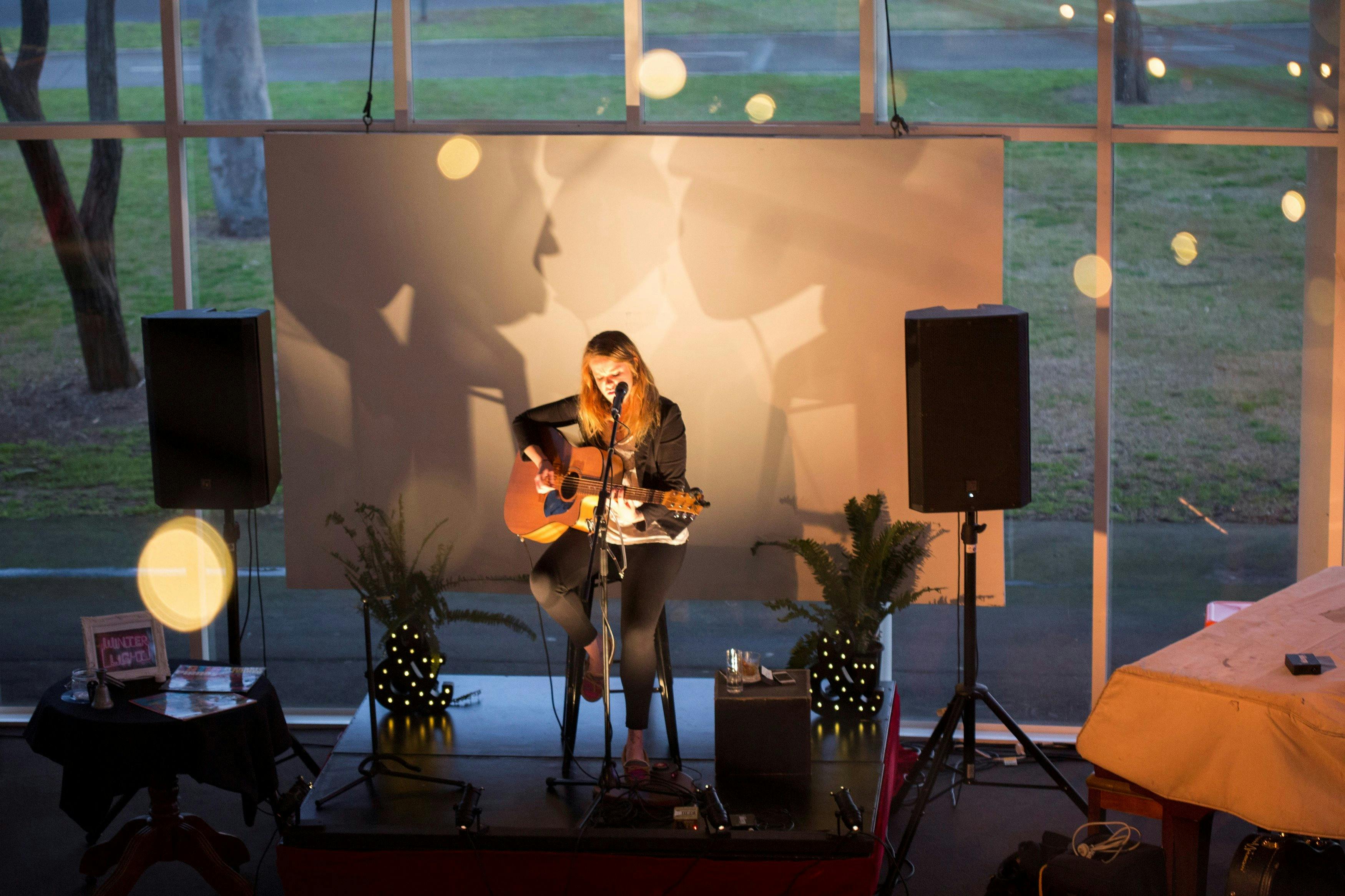 Woman playing a guitar on an informal stage in front of a large window