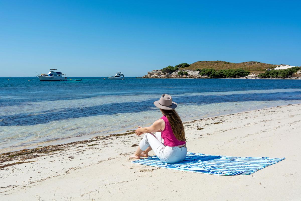 Woman sitting on a towel on a white sandy beach looking out over blue water.