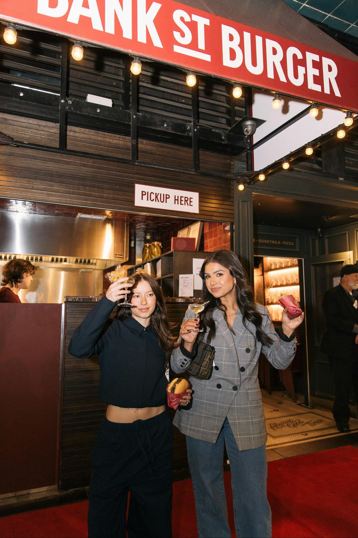 Two women stand outside Bank Street Burger raising their fresh burgers into the air giggling