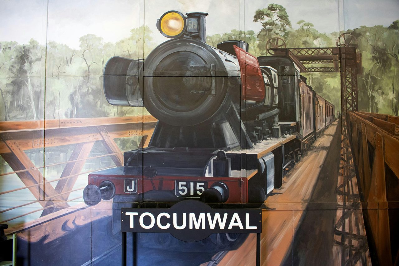 Mural of steam engine crossing bridge, with a railway station sign saying 'Tocumwal' in front.