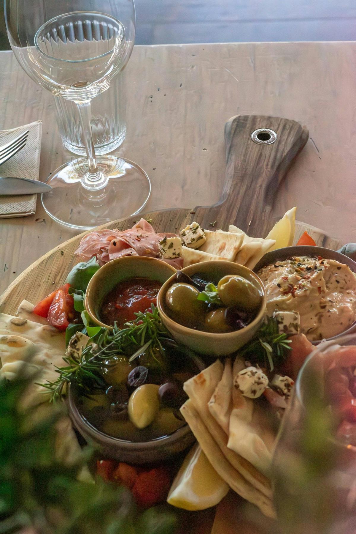 ooden antipasto board with olives, dips, cheeses, flatbread, and cold cuts beside wine glasses.