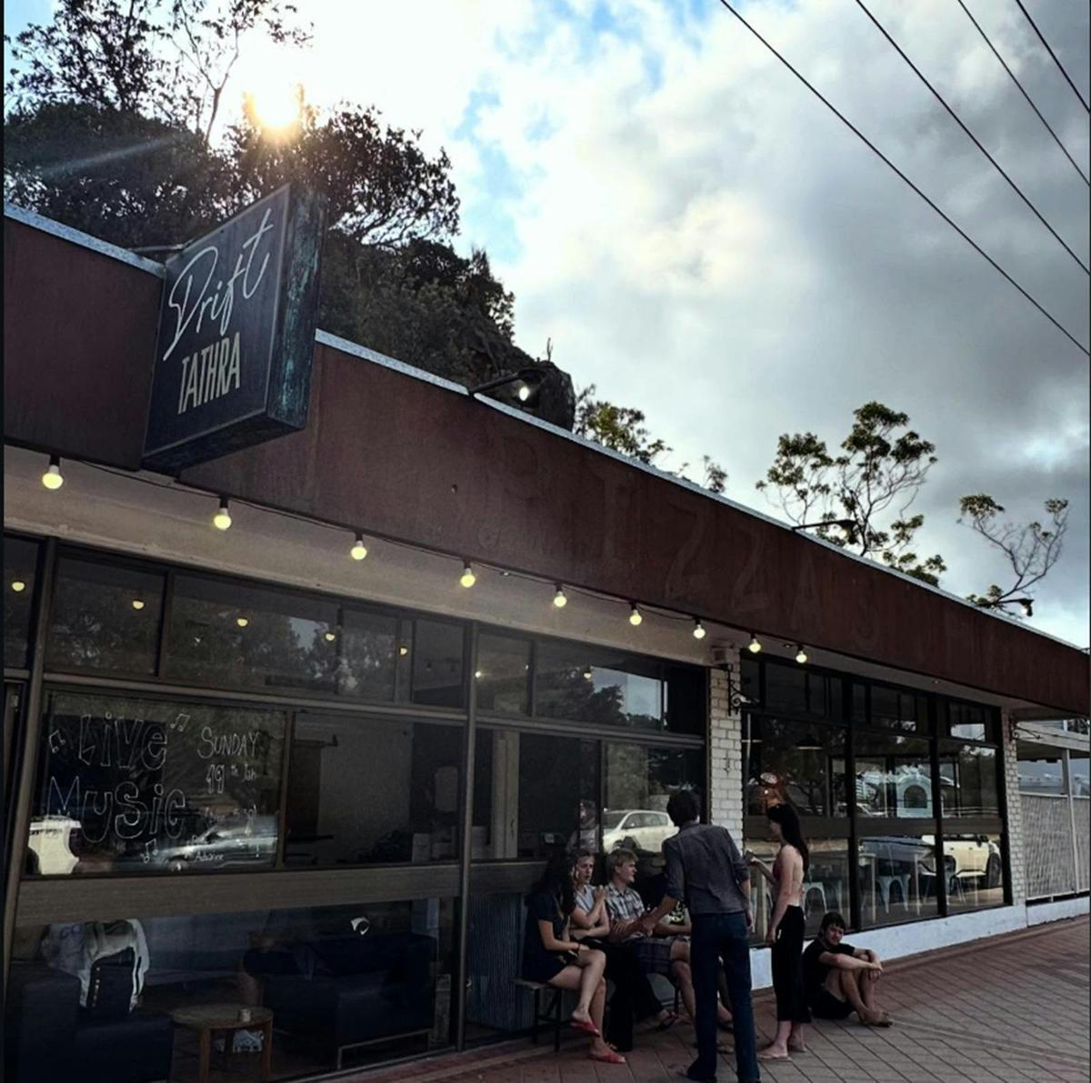 People gather outside the Drift Tathra pizza restaurant under string lights and a cloudy sky.