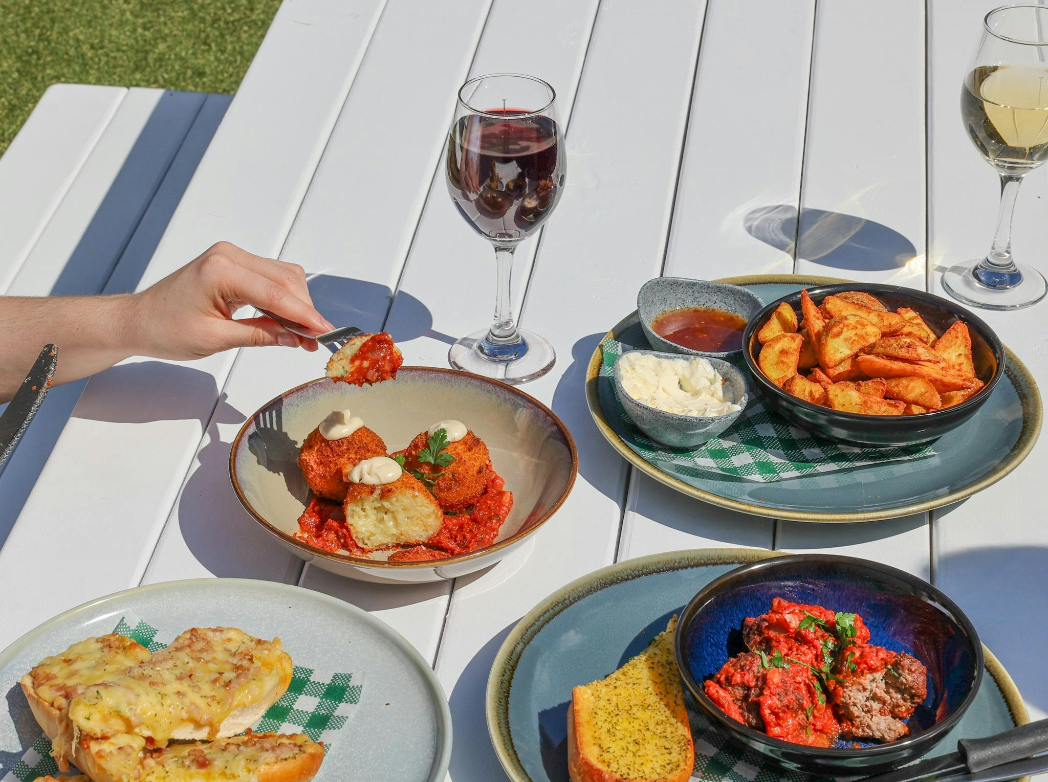 An array of food and wine on a picnic table including potato wedges, arancini balls, and bread