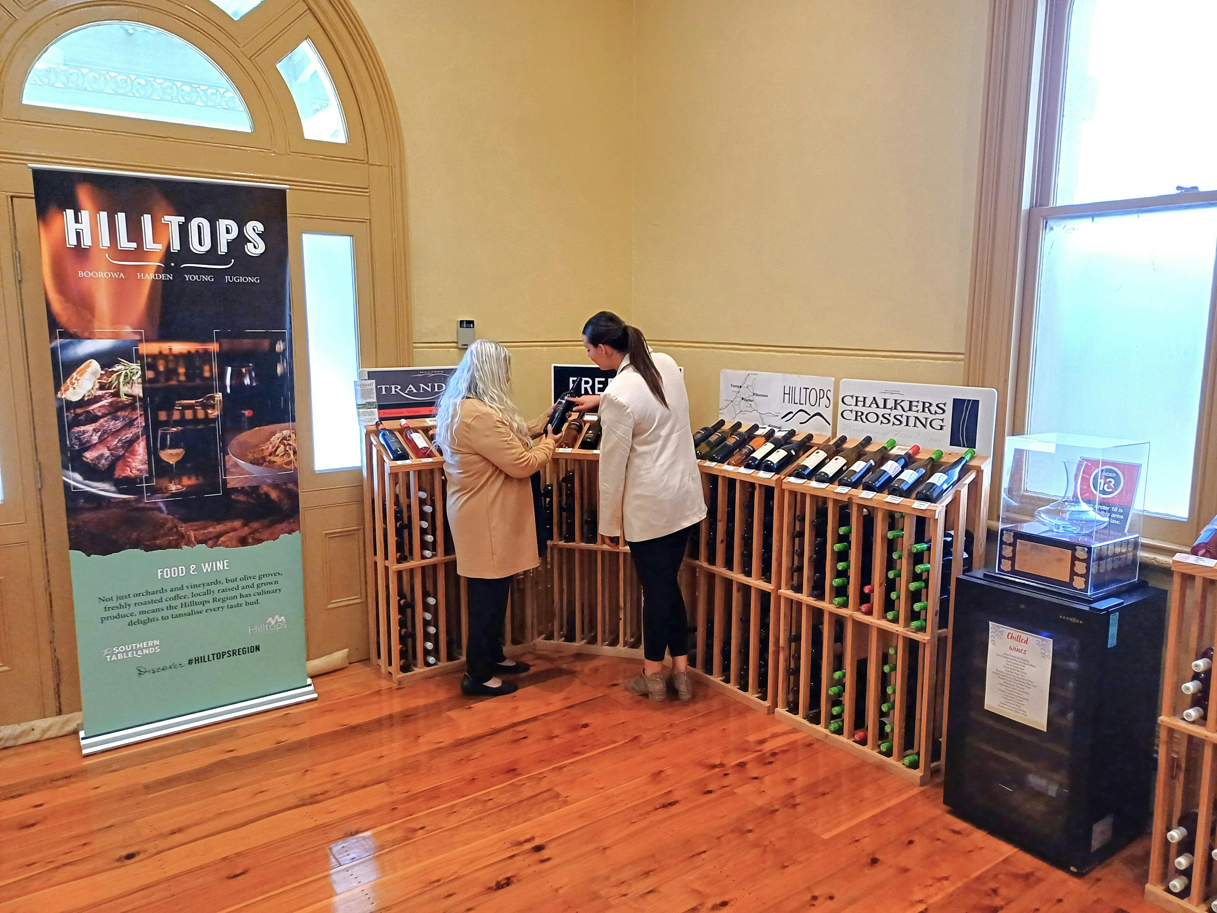 One corner of room with two women browsing wines. There is a Hilltops Wine banner and trophy too.