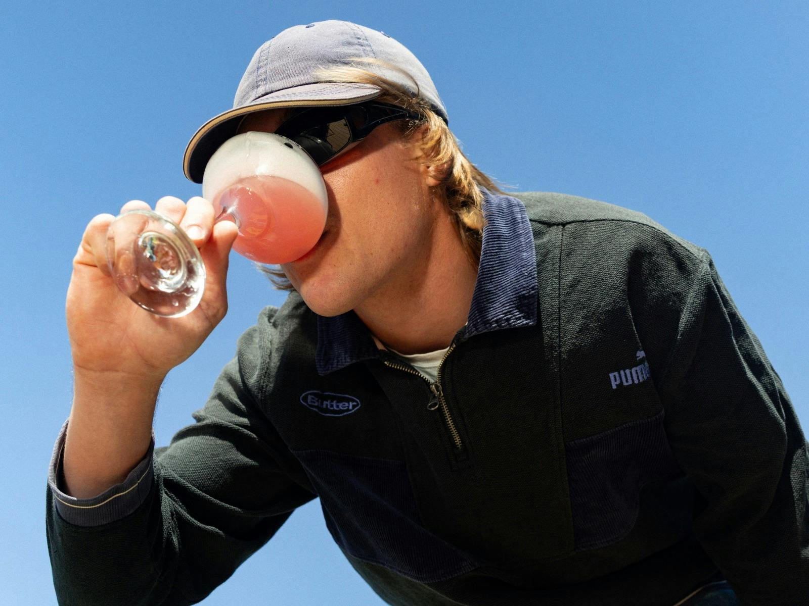 Man drinking a pink spritz cocktail outdoors against a clear blue sky.