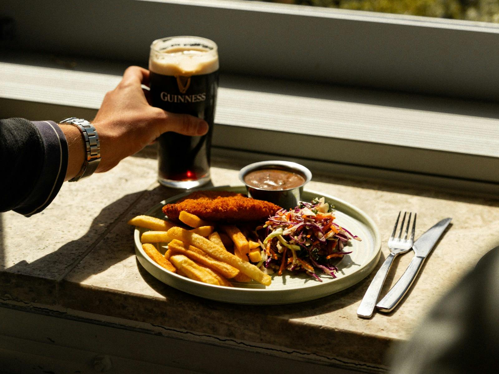 Hand holding a pint of Guinness with schnitzel, chips and slaw served on a plate.