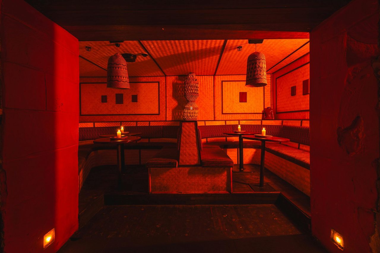Cozy nightclub booth with bamboo walls, red lighting, bench seating, candles, and woven lamps